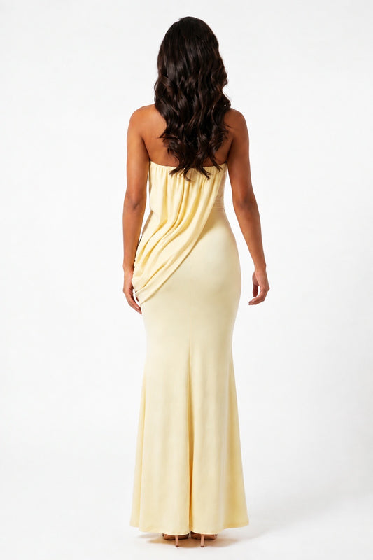 A woman viewed from the back wearing a strapless pale yellow maxi dress with draped detailing on the back, set against a plain white studio background.