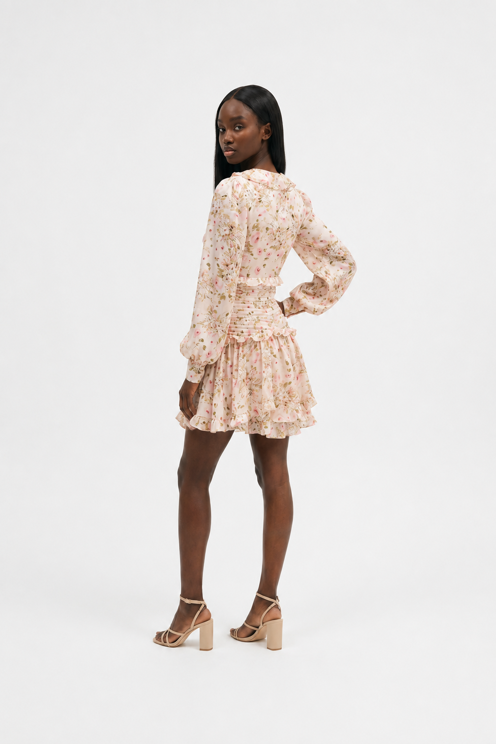 A woman modeling a long-sleeved mini dress with a delicate pink and green floral pattern on a cream base, shown from a three-quarters back view against a plain white studio background.