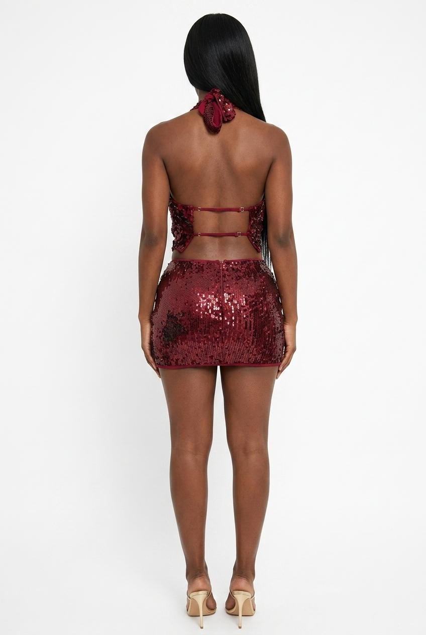 A woman shown from the back wearing a sparkling burgundy sequined halter mini dress with a matching fabric hair scrunchie, paired with beige high-heeled sandals, against a plain white studio background.