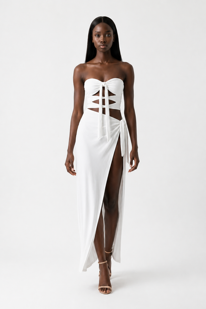 A woman modeling a strapless white maxi dress with a lace-up corset-style bodice, side cutouts, and a high front slit, photographed against a plain light gray studio background.
