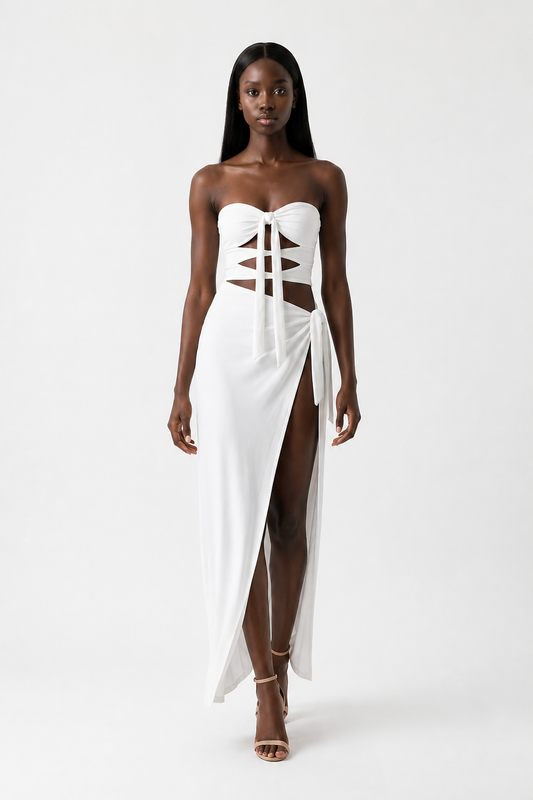 A woman modeling a strapless white maxi dress with a lace-up corset-style bodice, side cutouts, and a high front slit, photographed against a plain light gray studio background.