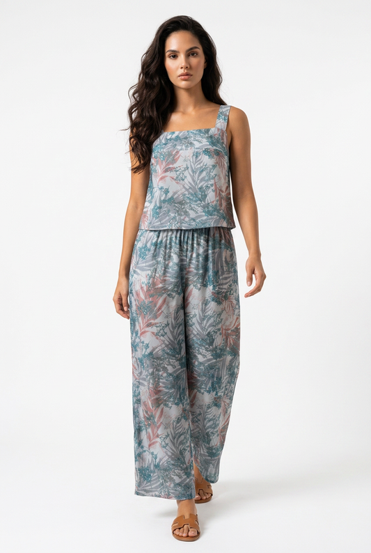 A woman modeling a sleeveless two-piece jumpsuit with a tropical leaf print in shades of teal, dusty blue, and muted pink, set against a plain white studio background.