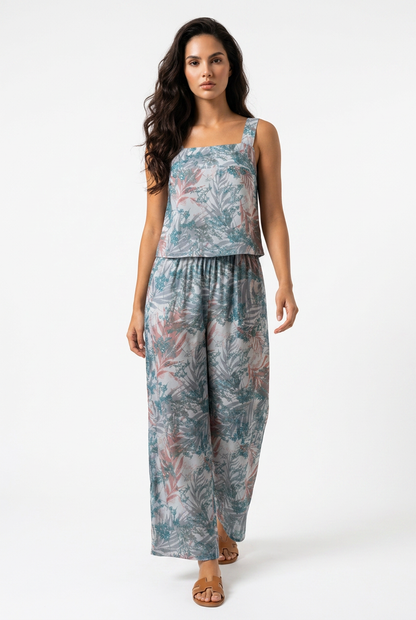 A woman modeling a sleeveless two-piece jumpsuit with a tropical leaf print in shades of teal, dusty blue, and muted pink, set against a plain white studio background.
