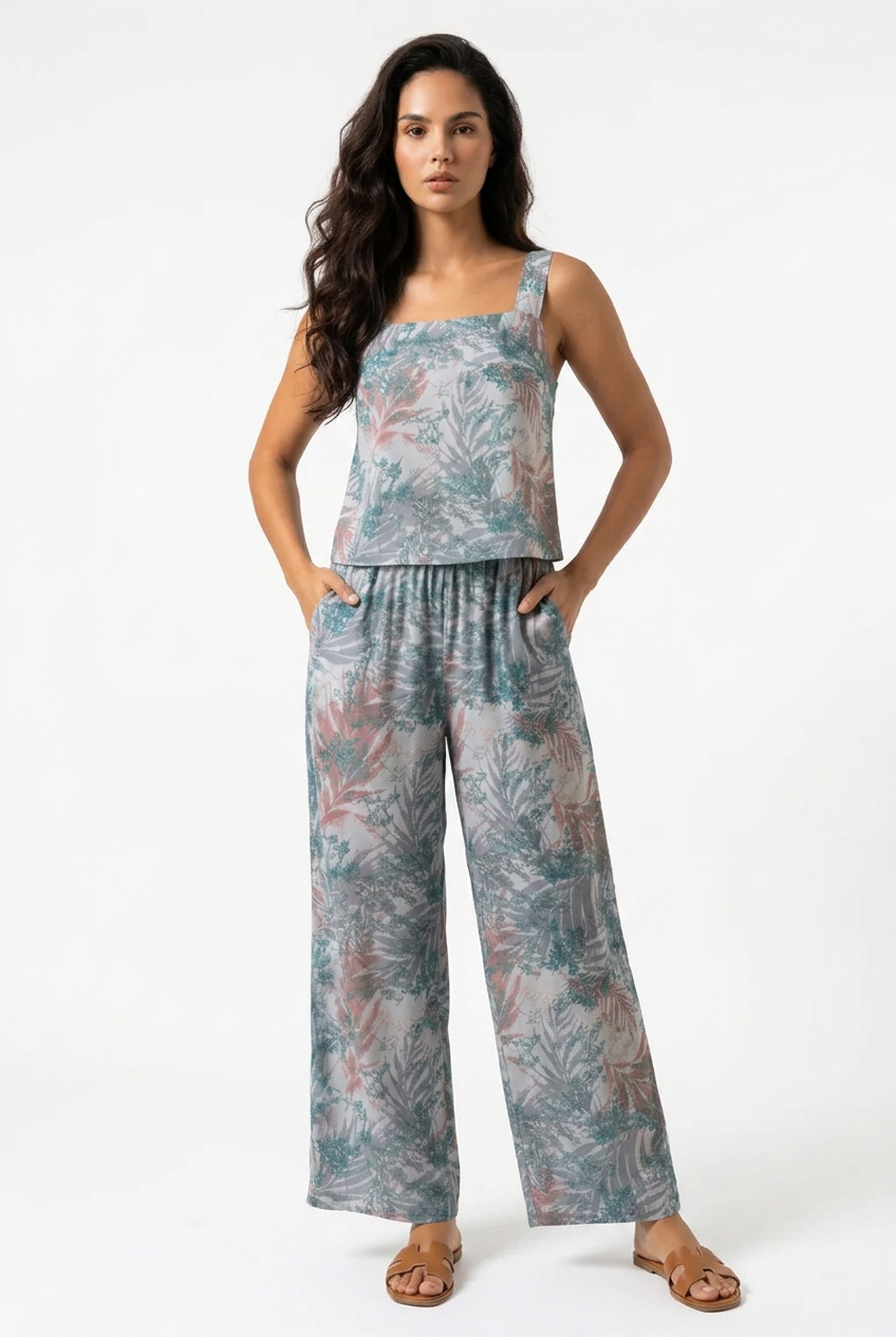 A woman modeling a sleeveless wide-leg jumpsuit with a tropical leaf print in muted teal, dusty pink, and gray tones, paired with brown flat sandals against a plain white studio background.