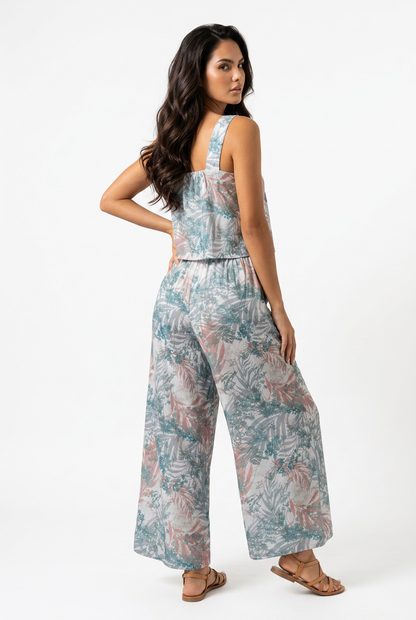 A woman modeling a sleeveless wide-leg jumpsuit with a tropical leaf print in shades of teal, dusty pink, and beige, shown from a three-quarters back view against a plain white studio background.