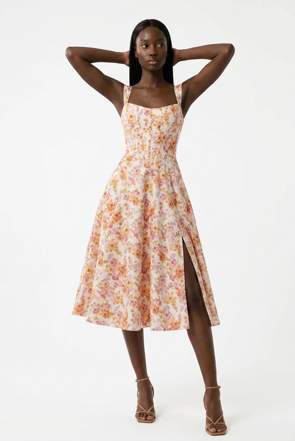 A woman modeling a sleeveless midi dress with a delicate orange and yellow floral print on a cream background, featuring thin straps, a smocked bodice, and a high side slit, set against a plain white studio background.