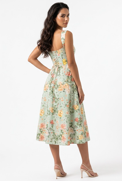 A woman modeling a sleeveless light green midi dress with a yellow, orange, and pink floral print, shown from a three-quarters back view against a white studio background.