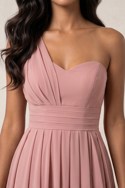 A woman wearing a dusty pink one-shoulder strapless dress with a sweetheart neckline, ruched bodice, and pleated skirt, shown against a neutral beige background.