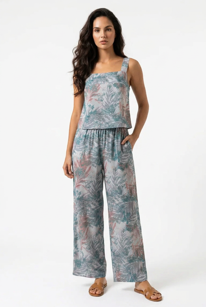 A woman modeling a sleeveless jumpsuit with a tropical leaf print in muted teal, blue, and dusty pink tones, paired with tan leather sandals, against a plain white studio background.