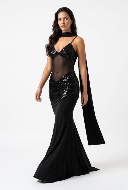 A woman modeling a black evening gown with a sheer mesh bodice, sequined embellishments, a high neck, and a long flowing cape-like drape, photographed against a plain light gray studio background.
