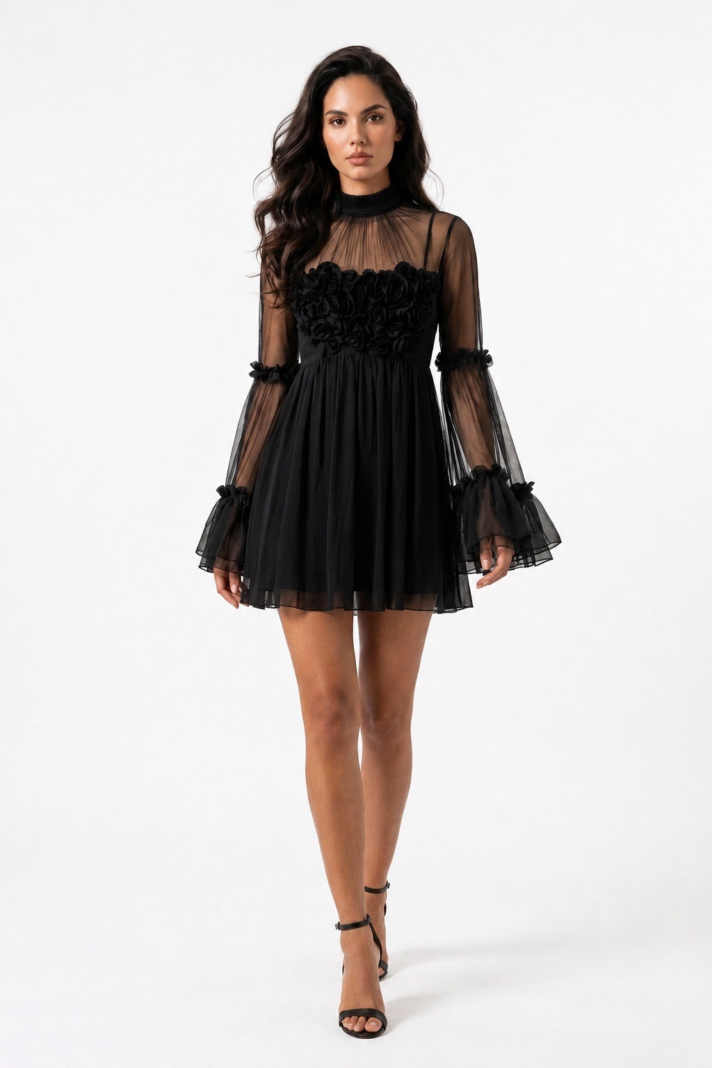 A woman modeling a short black tulle dress with a sheer high neckline, a black floral applique bodice, and dramatic flared sheer sleeves with ruffled trim, photographed against a plain white studio background.