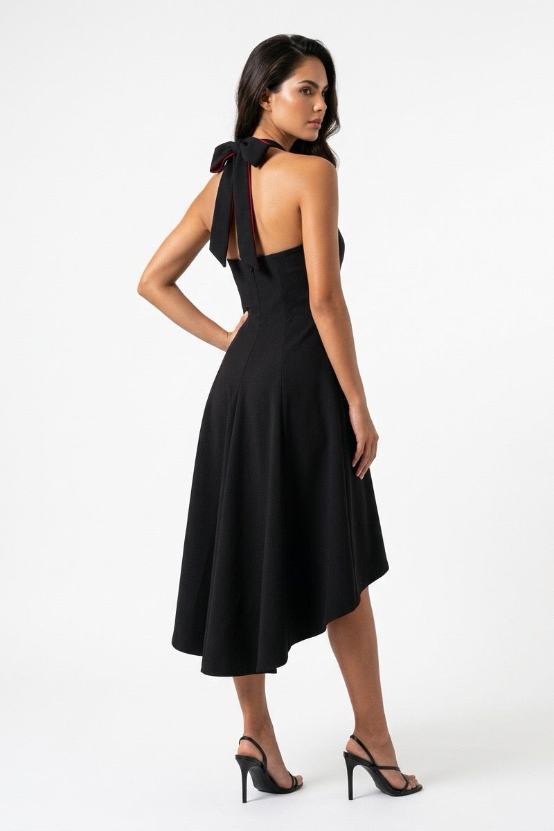 A woman modeling a black halter midi dress with a bow detail at the back of the neck, shown from a three-quarters back angle against a plain white studio background.