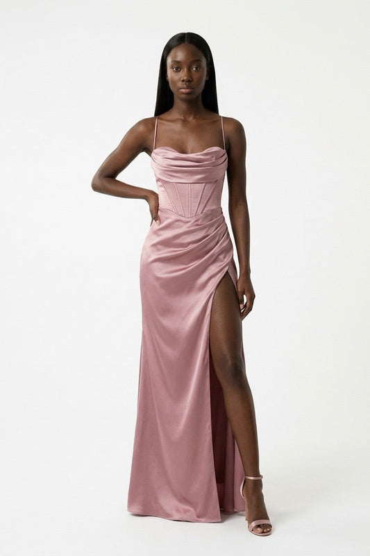 A woman modeling a floor-length dusty pink satin gown with spaghetti straps, a structured corset-style bodice, and a high thigh slit, photographed against a clean white studio background.