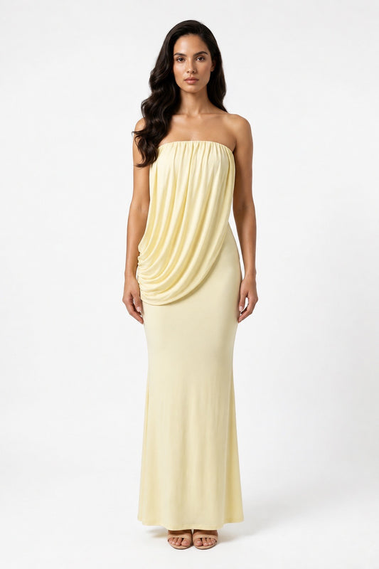 A woman modeling a strapless pale yellow maxi dress with draped detailing on the bodice and a fitted silhouette, photographed against a plain light gray studio background.
