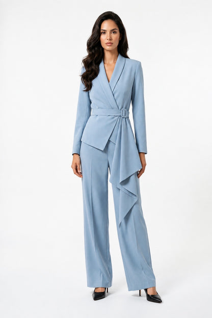 A woman modeling a light blue wrap-style jumpsuit with long sleeves, a matching waist tie, and wide-leg draped pants, photographed against a plain white studio background.