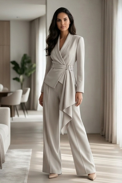 A woman stands in a bright, modern room wearing a light warm gray wrap-style jumpsuit with long sleeves, wide legs, and a draped asymmetrical hem detail.